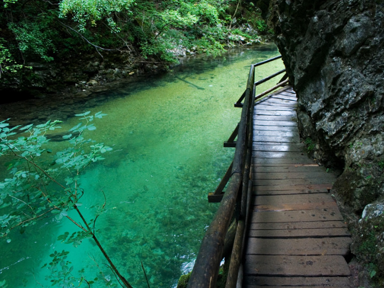 una passerella di legno addossata ad una parete di roccia costeggia un fiume dalle acque azzurrissime