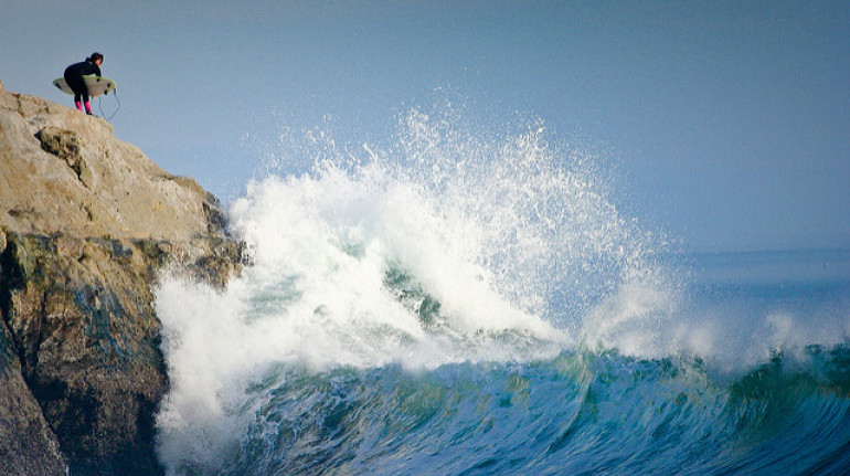 un'onda gigante si infrange su una scogliera, mentre un surfista la osserva dall'alto